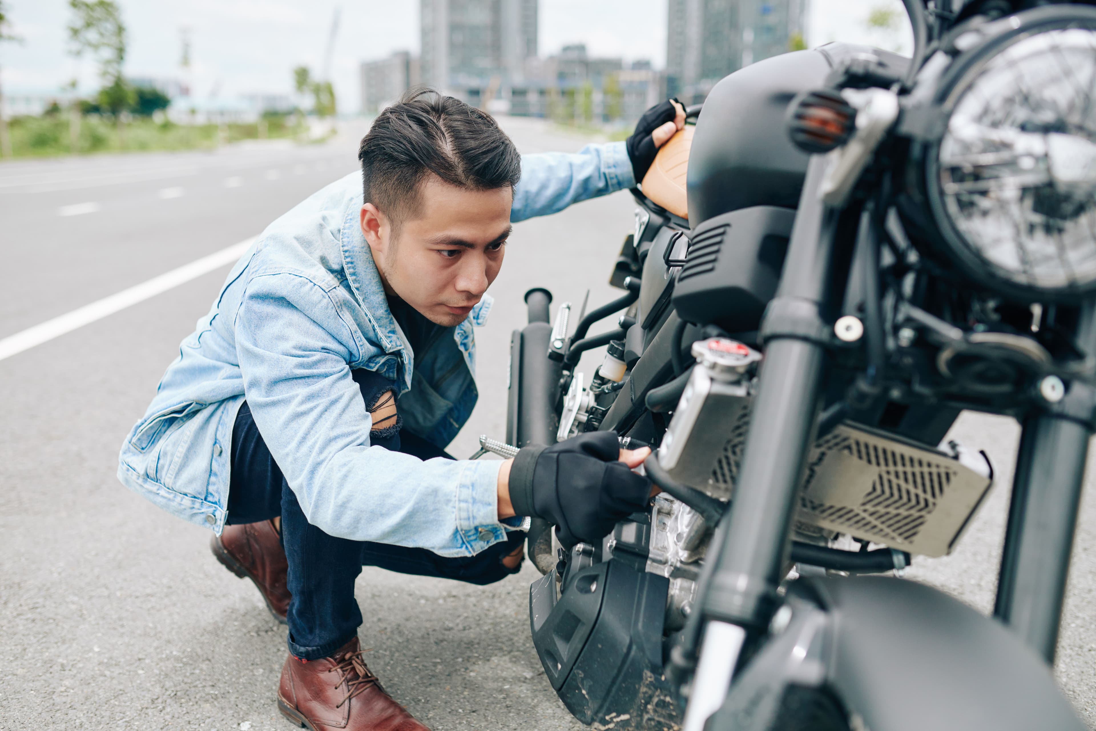 a man croutches beside a motorcycle on the side of the road, performing safety checks
